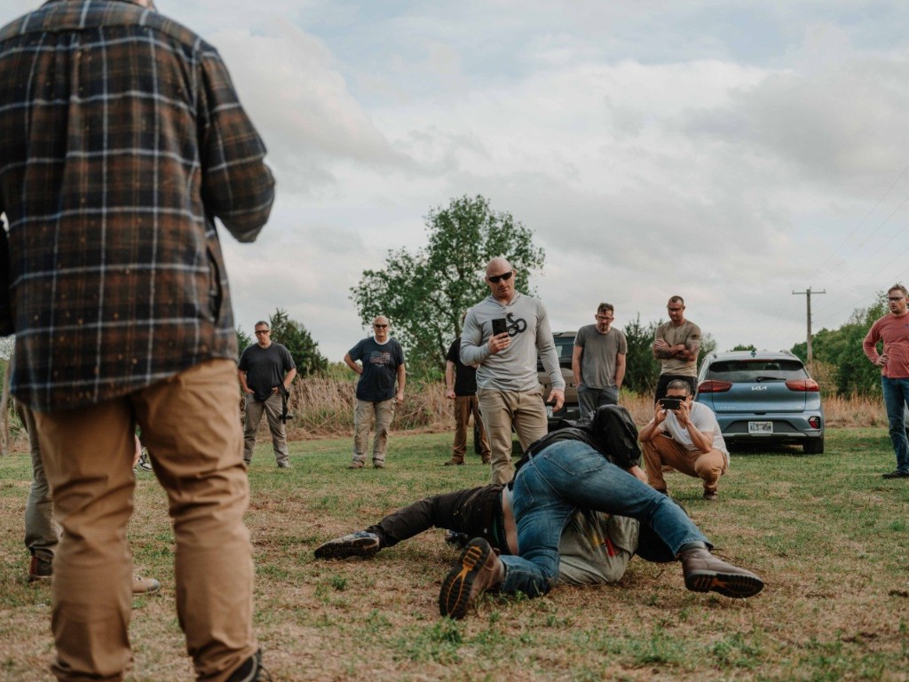 Students training grounded grappling while instructors observe on an outdoor range